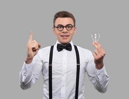 He has got an idea. Surprised young man in bow tie and suspenders holding a light bulb and gesturing while standing against grey background photo