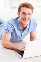 Surfing web at home. Handsome young man working on laptop and smiling while lying on the floor at his apartment photo