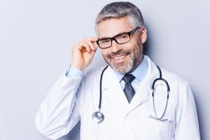 Experienced and confident doctor. Cheerful mature doctor adjusting his eyeglasses and smiling while standing against grey background photo