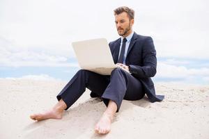 Businessman in desert. Confident young man in formalwear working on laptop while sitting on sand in desert photo