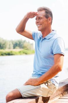 Spending time on quayside. Side view of cheerful mature man looking away and touching forehead with hand while sitting at the quayside photo