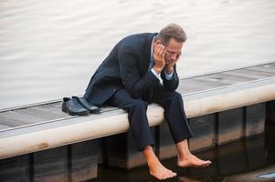 Depressed businessman. Depressed mature businessman holding head in hands while sitting barefoot at the quayside photo