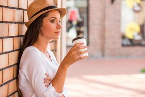 Relaxing with cup of fresh coffee. Side view of thoughtful young woman in funky hat holding cup with hot drink and looking away while leaning at the wall outdoors photo