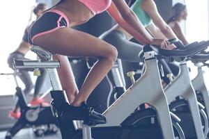 Power and motivation. Side view part of young women with perfect bodies in sportswear looking at camera with smile while cycling at gym photo