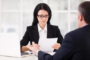 HR director and job candidate. Confident young woman in formalwear holding a paper and looking at it while grey hair man sitting in front of her and gesturing photo