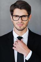 Ready to win. Confident young businessman adjusting his necktie and looking at camera while standing against grey background photo