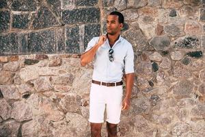 Staying cool and stylish. Handsome young African man in smart casual clothes adjusting his shirt collar while standing against the stoned wall outdoors photo