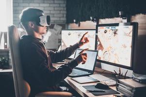 Testing games. Young man wearing virtual reality headset and gesturing while sitting at his desk in creative office photo