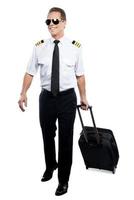 piloto alegre. piloto masculino confiado en uniforme caminando y llevando maleta mientras está aislado en fondo blanco foto