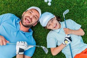 golfistas felices. vista superior de un niño alegre y su padre sosteniendo palos de golf y sonriendo mientras yacía en la hierba verde foto