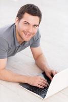 Spending his free time in the net. Top view of cheerful young man lying on the floor with laptop and looking at camera photo