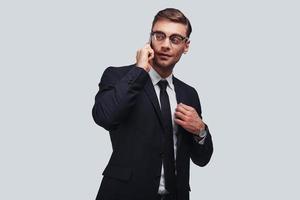 Good business talk. Handsome young man talking on smart phone and looking away with smile while standing against grey background photo
