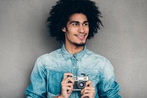 Modern photographer. Handsome young African man holding retro styled camera and looking away with smile while standing against grey background photo