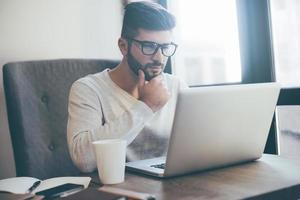 Thinking about opportunities. Thoughtful young man in glasses working on laptop and holding hand on chin while sitting in office or cafe photo