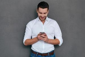 Texting business message. Handsome young man talking on the holding smart phone and looking at it with smile while standing against grey background photo