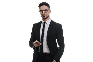 Confident and successful. Good looking young man in full suit holding smart phone and looking at camera while standing against white background photo