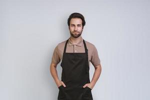 Handsome young man in apron looking at camera and holding hands on hip while standing against gray background photo