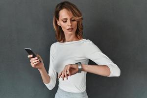 Time is money. Beautiful young woman in smart casual wear checking the time and holding a mobile phone while standing against grey background photo