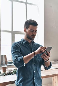 Checking messages from clients.  Handsome young man using smart phone and smiling while standing in the office photo