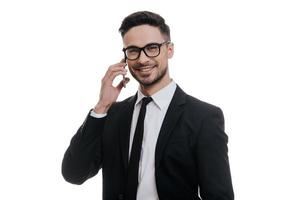 Good business talk. Handsome young man in full suit talking on smart phone and looking at camera while standing against white background photo