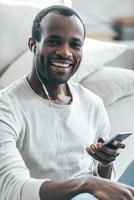 Listening a great song. Handsome young African man looking at camera and listening music while sitting on the carpet at home photo