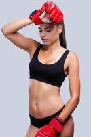Feeling tired after training. Attractive young sporty woman in boxing gloves touching her forehead and keeping eyes closed while standing against grey background photo