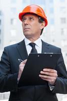 Confident foreman. Thoughtful mature man in formalwear and hardhat writing something in clipboard and looking away while standing outdoors photo