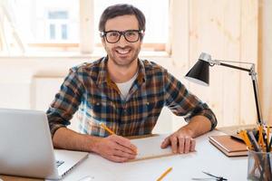 My space to create. Handsome young man designing project and smiling at camera while sitting at his working place photo