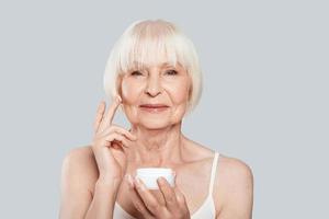 Anti aging. Beautiful senior woman applying beauty product while standing against grey background photo