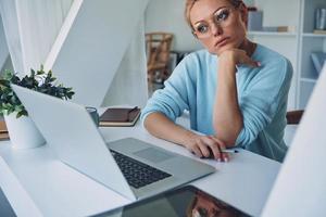 Beautiful young woman using laptop and looking thoughtful while working in office photo