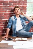 Good talk. Handsome young man sitting on the floor and talking on the mobile phone while digital tablet and documents laying near him photo