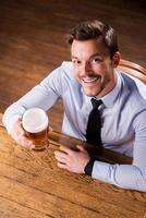 celebrando el éxito. vista superior de un apuesto joven con camisa y corbata sosteniendo un vaso con cerveza y sonriendo mientras se sienta en el mostrador del bar foto
