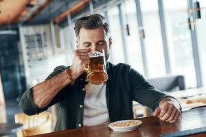 Charming young man in casual clothing drinking beer while spending time in the pub photo