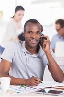 Young and creative. Handsome young African man talking on the mobile phone and smiling while people working on background photo