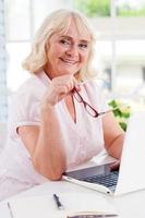 Feeling young and energetic. Happy senior woman using laptop and smiling at camera while sitting at the table photo