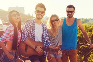 Having good time together. Group of happy young people bonding to each other and looking at camera while standing on the roofwith guitar photo