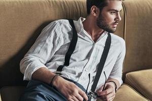 Handsome thinker. Handsome young man holding his eyewear and looking away while lying on sofa photo