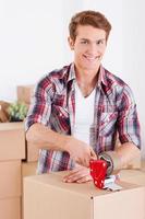 Getting ready for moving. Handsome young man packing boxes and smiling while other cardboard boxes laying in the background photo