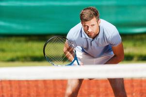 Confidence and full concentration. Handsome young man in polo shirt holding tennis racket and looking concentrated while standing on tennis court photo