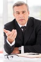 Tell me about yourself Confident mature man in formalwear looking at camera and gesturing while sitting at his working place photo