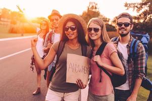 Going anywhere. Beautiful young people with backpacks hitchhiking on the road and looking happy photo