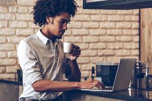 Checking all supplies. Side view of young African man using laptop and holding coffee cup while standing at bar counter photo