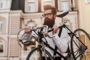 hombre barbudo con bicicleta retro. vista de ángulo bajo de un joven barbudo confiado que lleva su bicicleta en el hombro y mira hacia otro lado mientras camina al aire libre foto