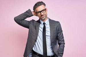 Relaxed mature man in formalwear holding hand behind head while standing against pink background photo