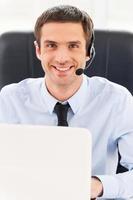 Always ready to help you. Cheerful young man in headset looking at camera while using laptop photo