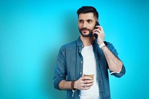 un joven apuesto con ropa informal hablando por teléfono inteligente mientras se enfrenta a un fondo azul foto