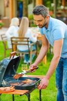 asar la carne a la perfección. joven confiado asando carne a la parrilla mientras otros miembros de la familia se sientan en la mesa del comedor en el fondo foto