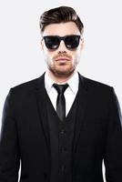 Elegance in style. Portrait of handsome young man in formalwear and sunglasses looking at camera while standing against grey background photo