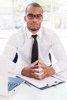 Confident businessman. Confident young African man in shirt and tie looking at camera while sitting at his working place photo