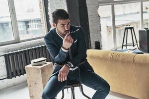 Magic look. Good looking young man in full suit putting on his eyewear while sitting on the stool photo